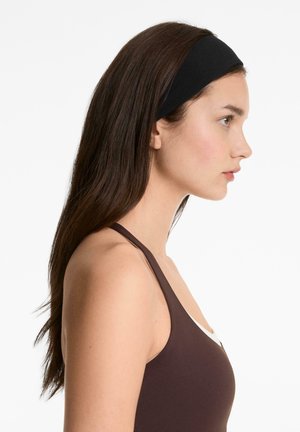 Bandă de cap elastică neagră purtată de un model, cu textură netedă, formă rotundă, expusă pe un fundal alb. Modelul are părul lung și brunet.