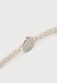 Ασημένιο ρολόι χειρός με μπρασελέ από ανοξείδωτο ατσάλι, στρογγυλή κάσα, λεία στεφάνη και κορώνα στο πλάι· η πίσω όψη διαθέτει εγχάρακτες λεπτομέρειες.