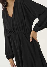 Robe noire avec un décolleté en V et des manches ballon, présentant des détails froncés à la taille et une jupe fluide. Texture lisse, tissu léger.