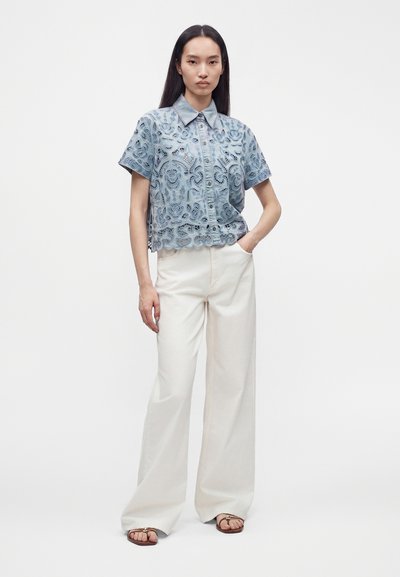 Femme debout, main dans la poche, portant une chemise en dentelle bleue à manches courtes, un pantalon blanc à jambes larges et des sandales ouvertes sur fond blanc.