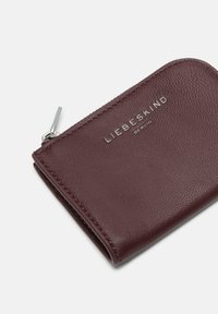 Bordowy skórzany portfel o fakturowanym wykończeniu, z wytłoczonym na przodzie srebrnym logo i srebrnym zamkiem błyskawicznym.