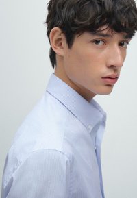 Camicia azzurra a righe sottili con colletto classico, caratterizzata da una leggera texture e dettagli con bottoni, indossata da un giovane con capelli mossi.