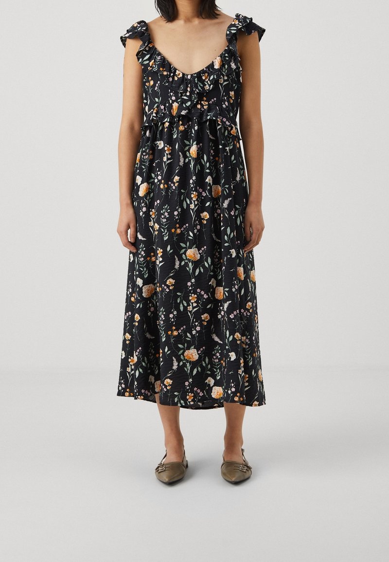 Blommig midiklänning i svart med volangkrage och ärmar, prydd med orange och rosa blommor på en strukturerad tyg. Stylad med platta skor.