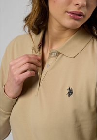 Beige polo shirt met een kraag, voorzien van twee knopen en een marineblauw logo op de linkerborst. Gladde stof met minimale textuur.