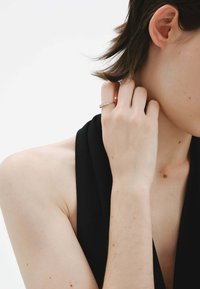 Anillo de oro con diamantes claros, llevado en una mano que toca el cuello. Prenda negra, textura de piel suave, con sutiles pecas visibles.