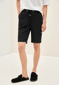 Svarta knälånga shorts i textil med elastisk midja och dragsko. I kombination med svarta halksandaler.