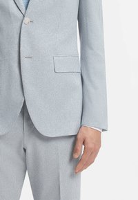 Veston de costume bleu clair à fines rayures avec pantalon assorti, porté avec une cravate à motifs, montrant le bras droit et la main détendus sur le côté.