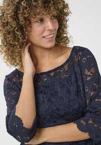 Top in pizzo blu navy con motivi floreali, maniche a tre quarti e scollatura ampia. Il tessuto presenta una trama trasparente e bordi a scalloped.