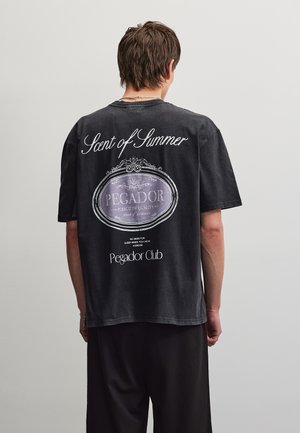 Személy fekete, túlméretezett pólót visel fehér felirattal "Scent of Summer" és "Pegador Club" a hátán, egyszínű háttér előtt.