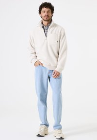 Fleece pullover in crèmekleur met een ritssluiting in de nek, een borstzak en geribde manchetten; gedragen met lichtblauwe jeans en beige sneakers.