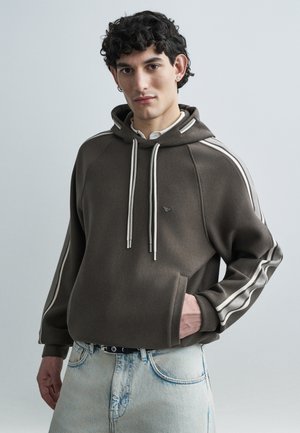 Bruine hoodie met witte zijstrepen, trekkoorden en voorzak; gecombineerd met lichtblauwe spijkerbroek. Gladde textuur en losse pasvorm.