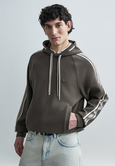 Sudadera con capucha marrón con franjas blancas en los lados, cordones y bolsillo frontal; combinada con vaqueros azul claro. Textura suave y ajuste relajado.