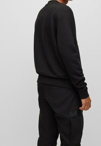 Sweat-shirt noir avec un col rond, des poignets et un ourlet côtelés. Le tissu semble doux avec une finition texturée. Porté avec un pantalon noir assorti.