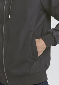 Sort bomberjakke med en glat, skinnende overflade; har en lynlås foran, ribbede manchetter og sidelommer. Diskret logoetikette ved kanten.