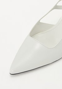 Chaussure blanche à bout pointu en cuir texturé, présentant des côtés découpés et une semelle noire. Les accents incluent des détails de couture minimalistes.