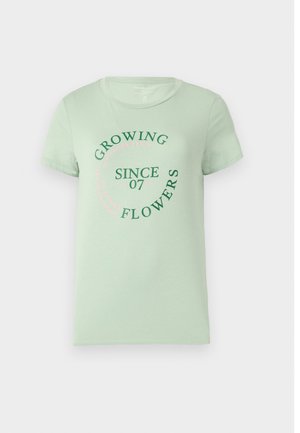 Camisa de algodón verde claro con mangas cortas. En el frente presenta un texto circular: "Cultivando Flores Desde 07" en verde y rosa. Con cuello redondeado.