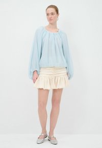 Helles blaues Blusenshirt mit Puffärmeln und gerafftem Ausschnitt; cremefarbener, rüschierter Minirock; Trägerin in silbernen Ballerinas mit Knöchelriemen.