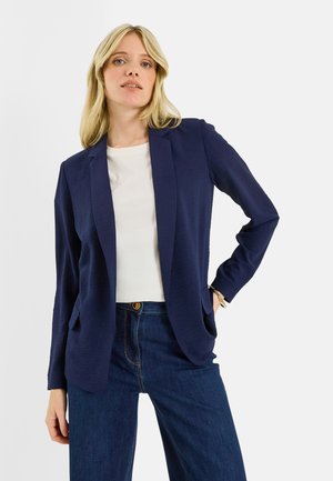 Femme blonde portant un blazer bleu marine, un haut blanc et un jean bleu, debout avec une main dans la poche devant un fond clair uni.