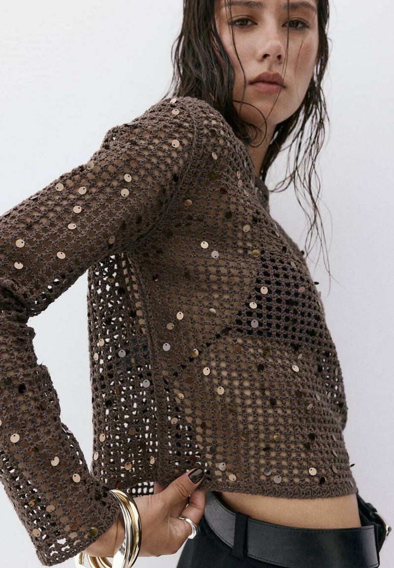Femme aux cheveux mouillés portant un pull en crochet marron avec des sequins, un haut noir en dessous, un pantalon noir et des bracelets en or à son poignet.