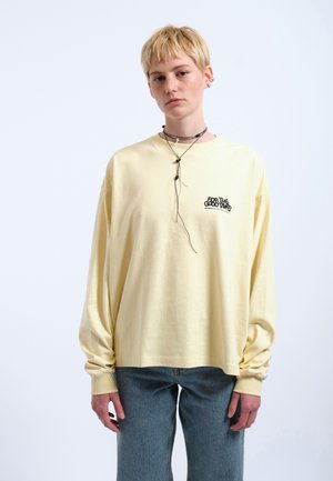 Młoda osoba z krótkimi blond włosami nosi oversize bladożółtą koszulę z długim rękawem z małym czarnym napisem, niebieskie dżinsy i kilkupoziomowe czarne naszyjniki.