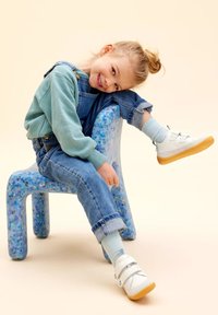 Bambino con salopette di denim blu e felpa verde menta, seduto su una sedia blu testurizzata, indossa scarpe bianche e calzini azzurri.