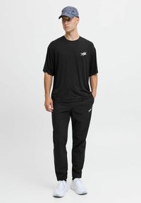 Camiseta negra de manga corta con un logo blanco, combinada con pantalones negros. El modelo lleva una gorra azul con estampado y zapatillas blancas.
