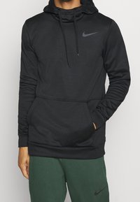 Homme portant un hoodie noir Nike avec une poche avant et des cordons, associé à un pantalon Nike vert foncé, se tenant devant un fond uni.