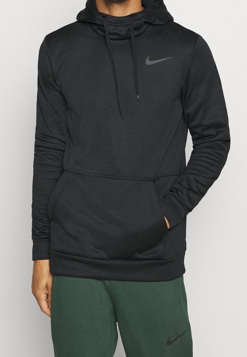 Homme portant un hoodie noir Nike avec une poche avant et des cordons, associé à un pantalon Nike vert foncé, se tenant devant un fond uni.