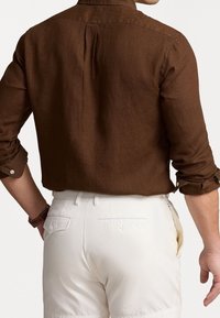 Uomo che indossa una camicia marrone a maniche lunghe con i polsini arrotolati e pantaloni bianchi con bottoni, visto da dietro su uno sfondo neutro.