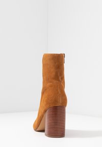 Bota de tobillo de ante marrón con pierna estrechada, cremallera lateral y un tacón de madera acanalado de 3 pulgadas, con una textura suave y mate.