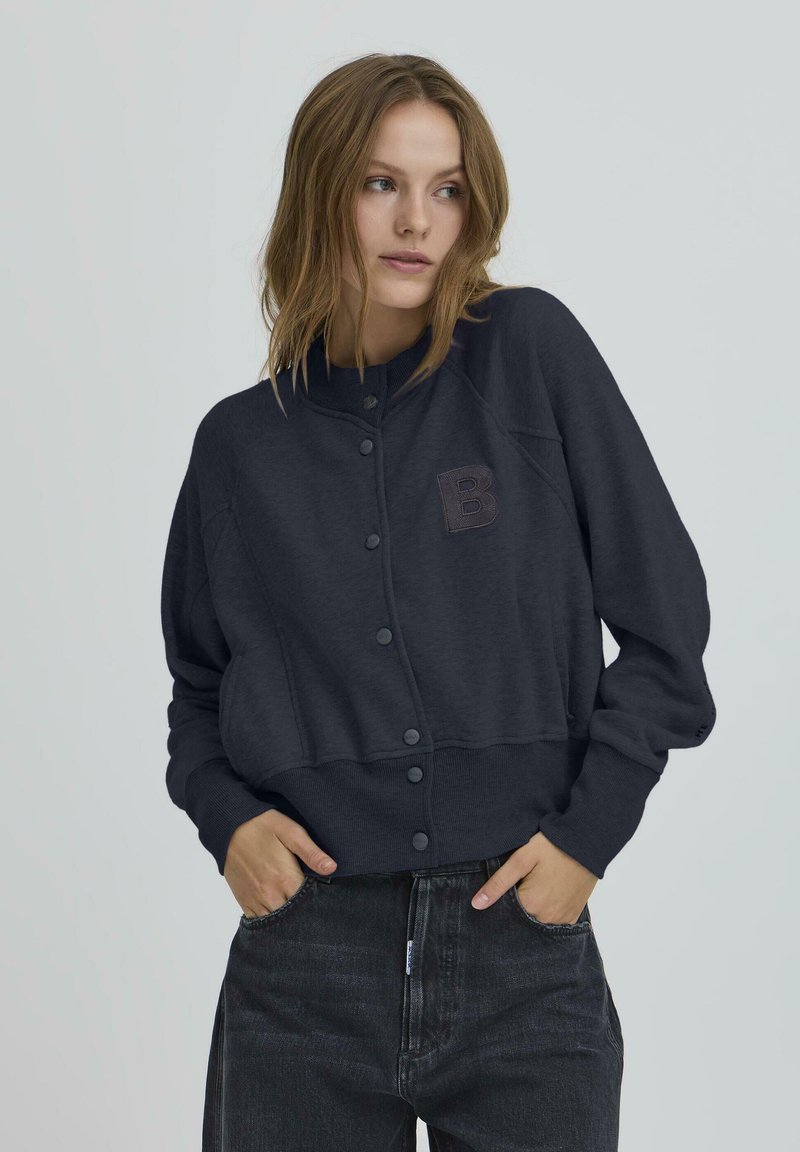 Tamsiai pilkas trumpas megztinis su apvaliu apykakle, mygtukų uždarymu ir tekstūruotu raide "B" akcentu. Pagamintas iš minkštos medžiagos, turi prigludusias rankoves.