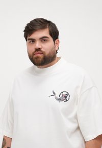 White short-sleeve T-shirt featuring a colorful fish graphic on the left chest. Made of smooth fabric with a loose fit and high collar.