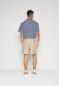 Kortärmad, blå mönstrad skjorta med geometrisk design, matchad med beige shorts. Vita sneakers med gröna och bruna detaljer.
