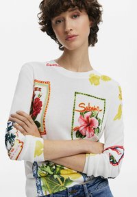 White sweater featuring colorful floral and textual patterns, round neckline, ribbed cuffs, and multi-colored accent designs.