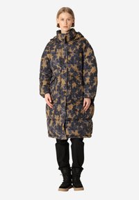 Abrigo tipo puff en patrón floral azul marino y beige, con capucha, cierre de cremallera y bolsillos laterales, usado sobre pantalones negros y botas.