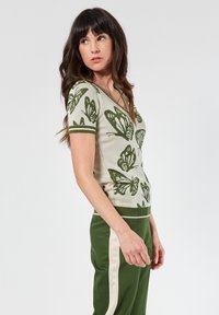 Top de punto con un patrón de mariposas verdes sobre un fondo beige claro, con escote en V y ribetes verdes en las mangas y el dobladillo.