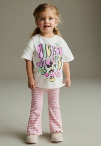 Weißes Baumwoll-T-Shirt mit buntem "Mickey Mouse"-Grafik, kombiniert mit rosa und weißen gestreiften, weit geschnittenen Leggings. Helle Schuhe mit Klettverschlüssen.