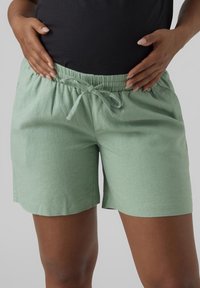 Personne portant un short à cordon de serrage vert clair et un haut noir, les mains reposant sur les hanches, visible de la taille jusqu'au milieu de la cuisse.