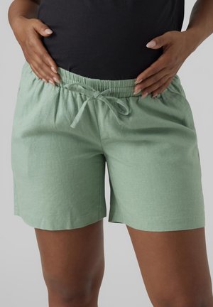 Person wearing light green drawstring shorts and a black top, hands resting on hips, visible from waist to mid-thigh.