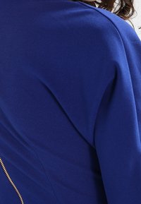 Top de tela azul royal con una textura suave y elegante, con un detalle de cremallera dorada visible en la parte posterior para acentuar.