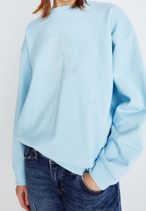 Personne portant un sweat-shirt bleu clair avec un motif de champignon en strass et un texte, associé à un jean bleu, main dans la poche, cadré au niveau du cou.