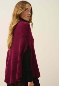 Burgundy ribbatstrikkad poncho med lös passform, sidöppningar och hög krage. Bärs över en svart långärmad topp, vilket visar på en texturkontrast.