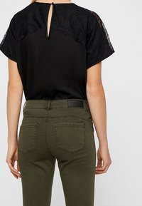 Top nero con maniche in pizzo e dettaglio a chiave sul retro, abbinato a pantaloni verde oliva con tasche posteriori e un'etichetta nera singola.