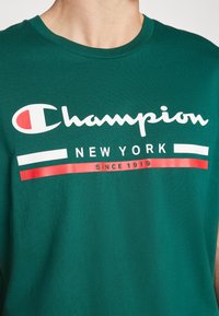 Camisa verde con el logo de Champion en blanco y rojo, el texto "NEW YORK" y "SINCE 1919" debajo del logo en el pecho.
