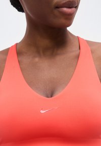 Γυναίκα που φοράει κοραλλί αθλητικό σουτιέν με εμφανές λογότυπο της Nike στο στήθος, εμφανίζοντας τον λαιμό και το κάτω μέρος του προσώπου της με φόντο μονόχρωμο.