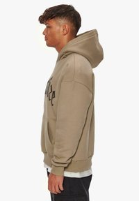 Hellbeige Kapuzenpullover mit Kängurutasche, oversized Schnitt und schwarzer Schrift auf der Vorderseite. Mit einer Kapuze mit Kordelzug und gerippten Bündchen.