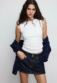 Top bianco aderente senza maniche abbinato a una gonna in denim scuro con due bottoni, tasche frontali e cuciture a contrasto, completato da un cardigan blu navy.