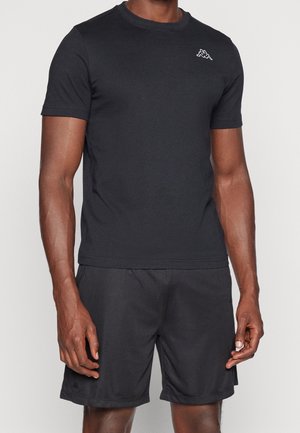 Homme portant un T-shirt noir à manches courtes et un short noir, debout avec les bras détendus le long du corps sur un fond clair uni.