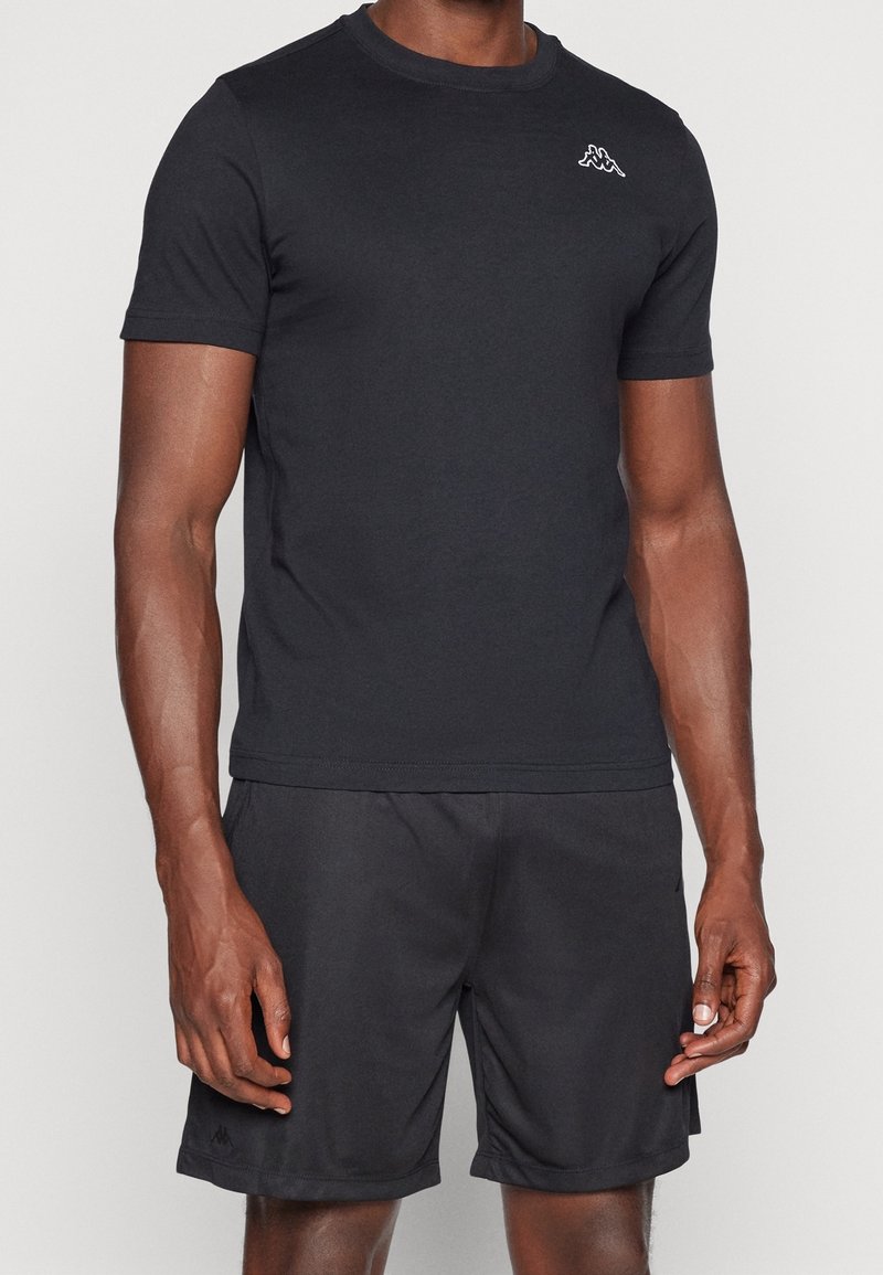 Homme portant un T-shirt noir à manches courtes et un short noir, debout avec les bras détendus le long du corps sur un fond clair uni.
