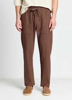 Uomo che indossa pantaloni marroni con coulisse e tasche frontali e scarpe slip-on color cuoio, in piedi davanti a uno sfondo bianco semplice.